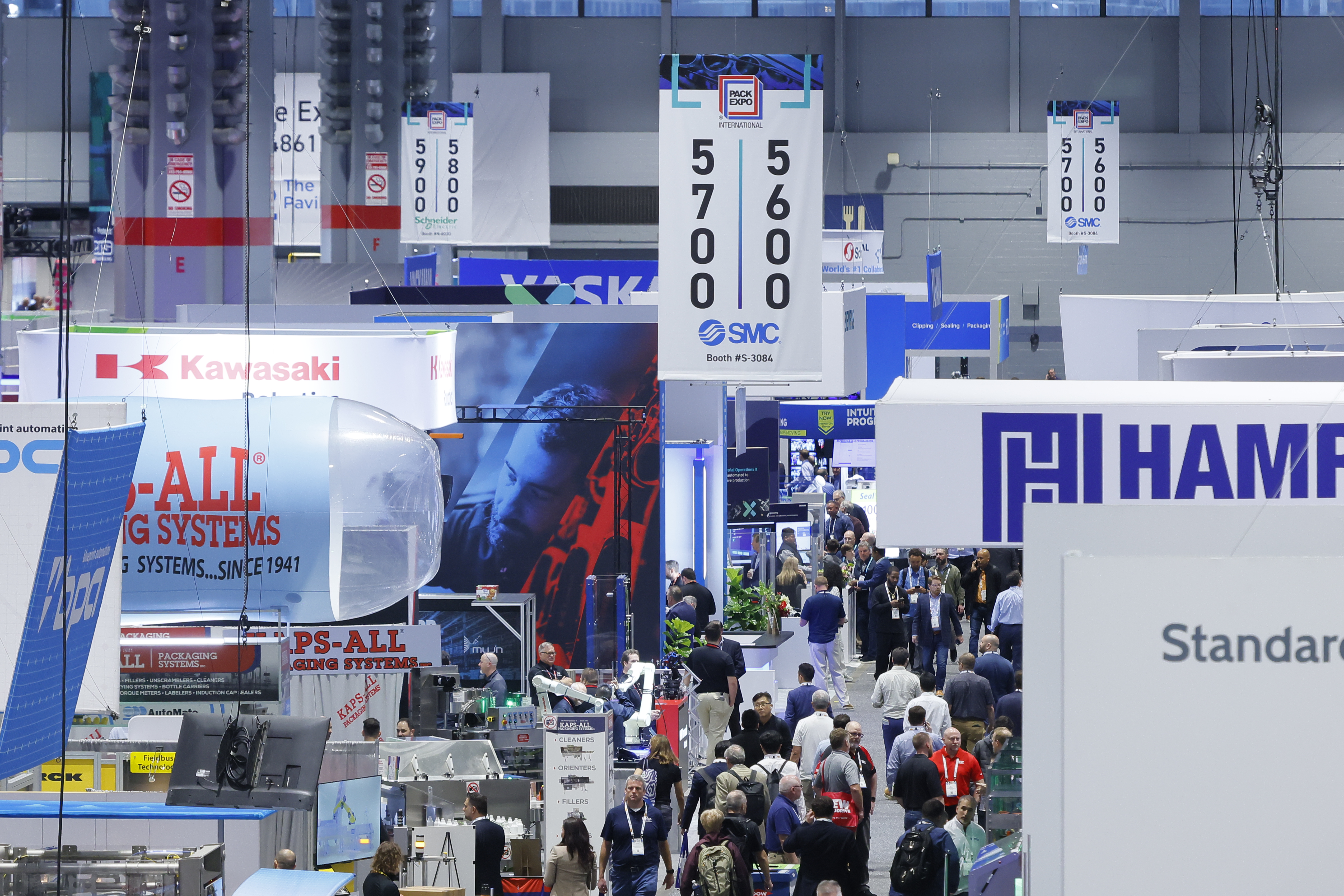 Recinto de una feria industrial abarrotado, con numerosos stands de empresas que exhiben carteles con los colores azul y rojo de sus marcas, así como pantallas digitales.